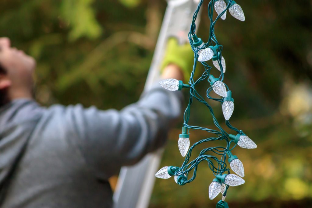 Man in gray sweatshirt climbing ladder to put up Christmas lights.