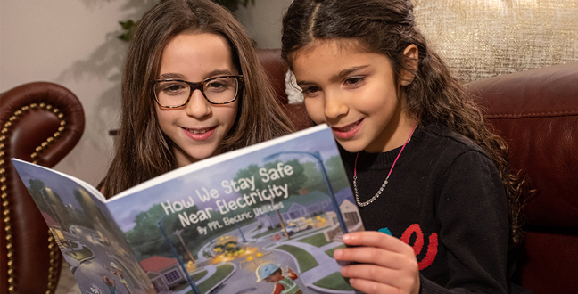 Two young girls sitting on couch reading How We Stay Safe Near Electricity Book/
