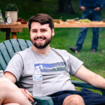 Man with brown hair and moustache and beard sitting in green lawn chair with bottled water in holder