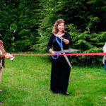 Two women holding red ribbon and woman in black with large scissors cutting ribbon