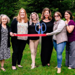 Six women posing with red ribbon and scissors