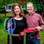 Woman in black dress and man in red button down shirt and jeans holding red ribbon and large scissors