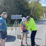 Man with hat, tie-dyed shirt and shorts talking with man in neon yellow sweatshirt and baseball cap next to video camera