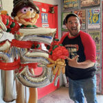 Man with red and black t-shirt with beret pointing to life sized balloon character holding a 75