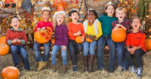 A group of young children sit in a row on stacked hay bales holding pumpkins. They wear bright clothes and make silly faces.