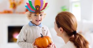 A child wears a head band with construction paper turkey on it. In front of him, an adult woman kneels down and hands him a sparkly orange pumpkin.