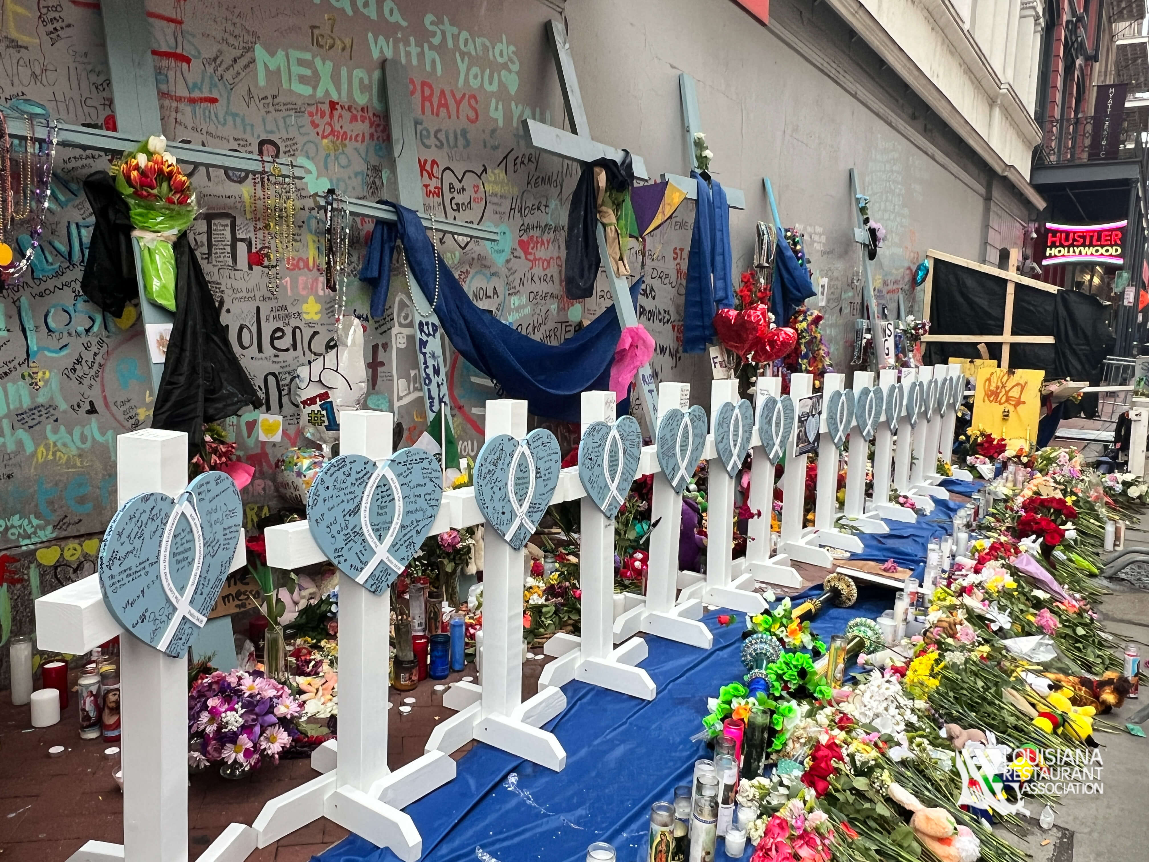 A memorial site with white crosses, blue hearts, flowers, candles, and written messages on a wall, honoring victims.