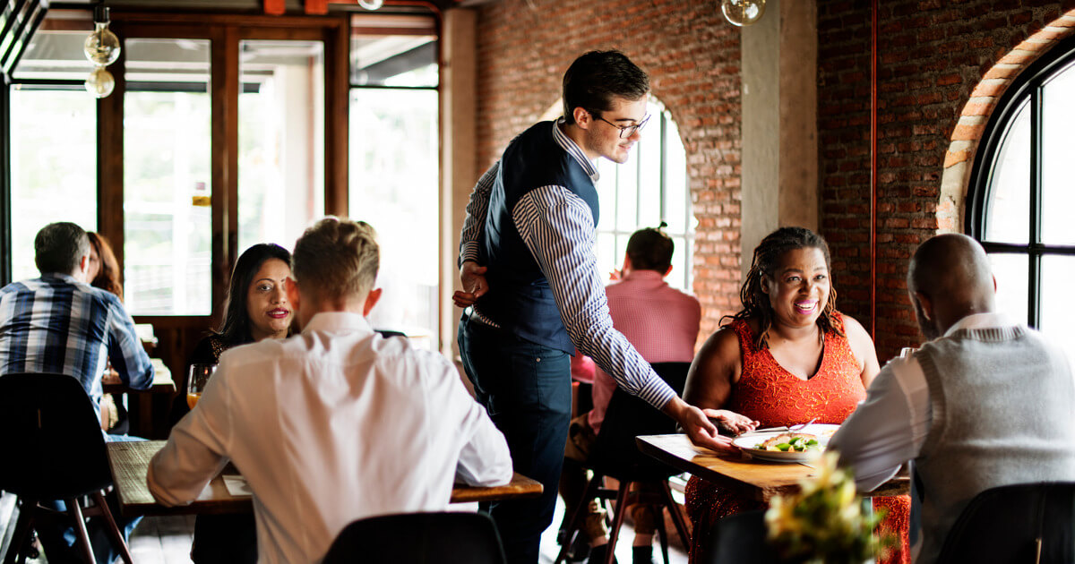 Server placing a dish on a table in a warmly lit restaurant with exposed brick walls and large windows.