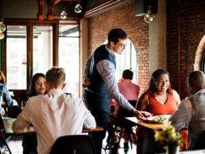 Server placing a dish on a table in a warmly lit restaurant with exposed brick walls and large windows.