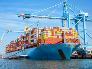 Cargo ship loaded with colorful containers at a port, with cranes and a wind turbine nearby.