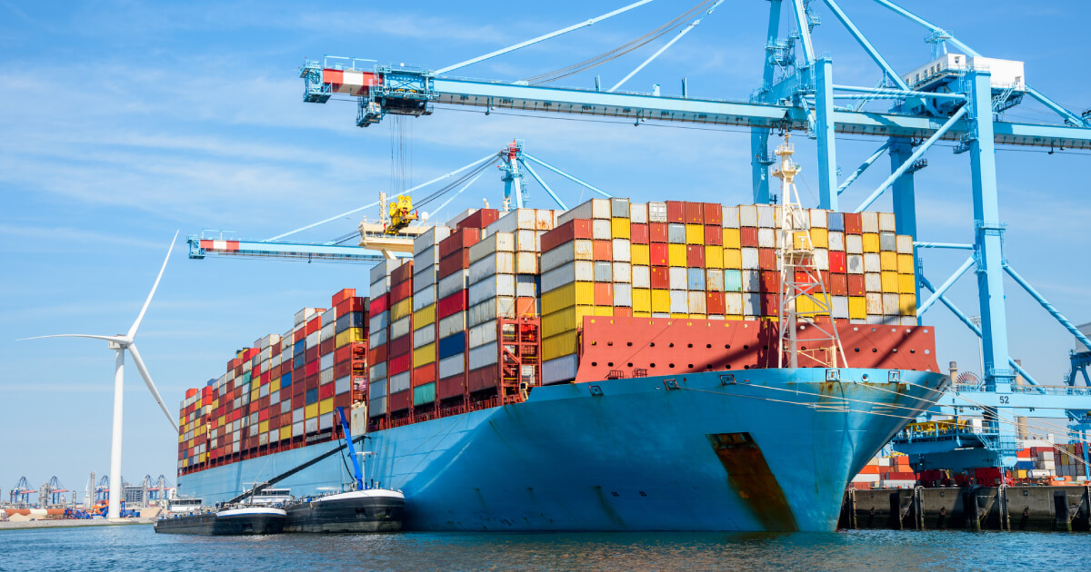 Cargo ship loaded with colorful containers at a port, with cranes and a wind turbine nearby.