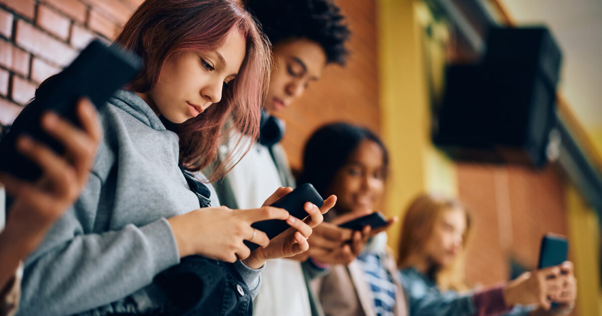 A group of young people standing in a line, each focused on their smartphones, immersed in their devices against a brick wall backdrop.