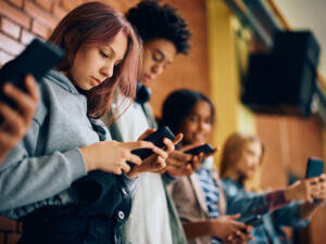 A group of young people standing in a line, each focused on their smartphones, immersed in their devices against a brick wall backdrop.