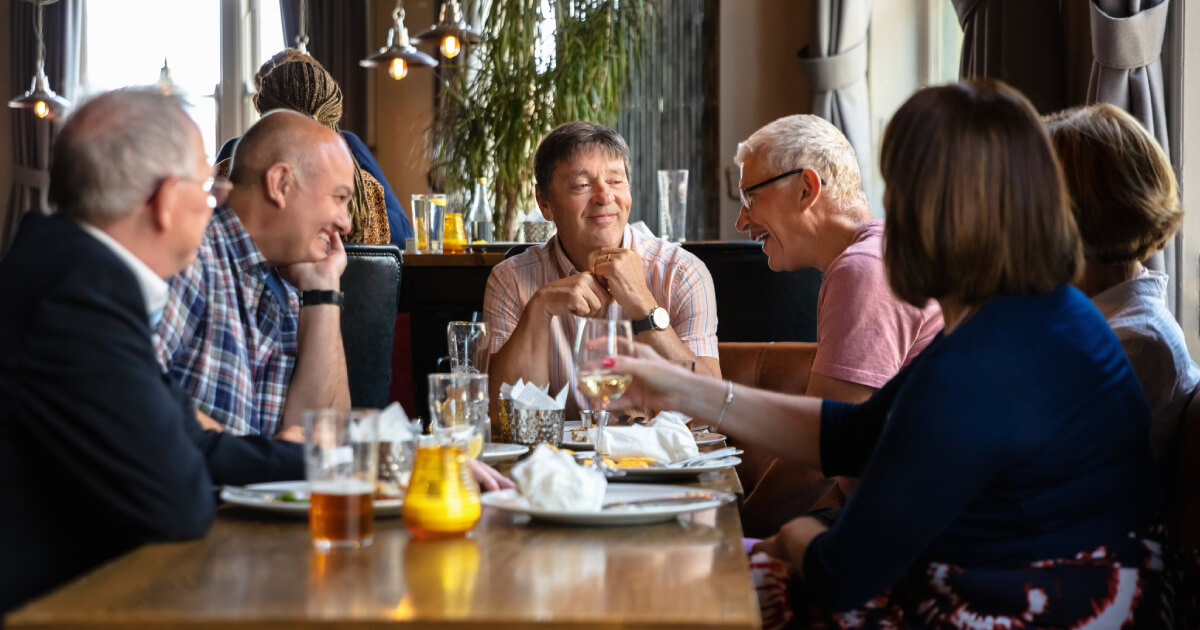 A group of six adults gather around a wooden table in a cozy restaurant, enjoying drinks and food while engaged in conversation.