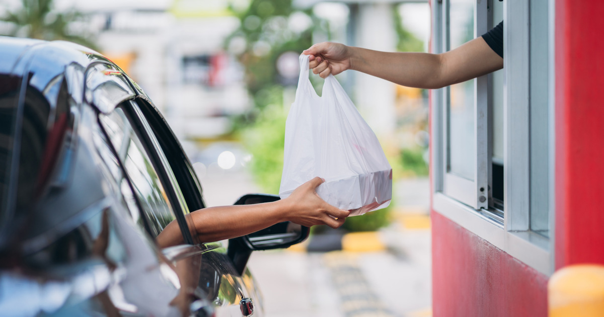 A drive-thru transaction with a person in a car receiving a plastic bag from a window in a red building.