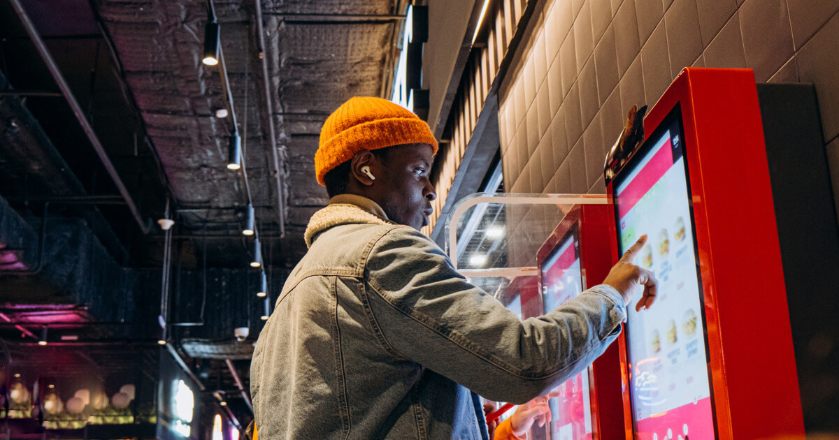 Person in an orange cap using a touchscreen kiosk in a dimly lit, modern setting.