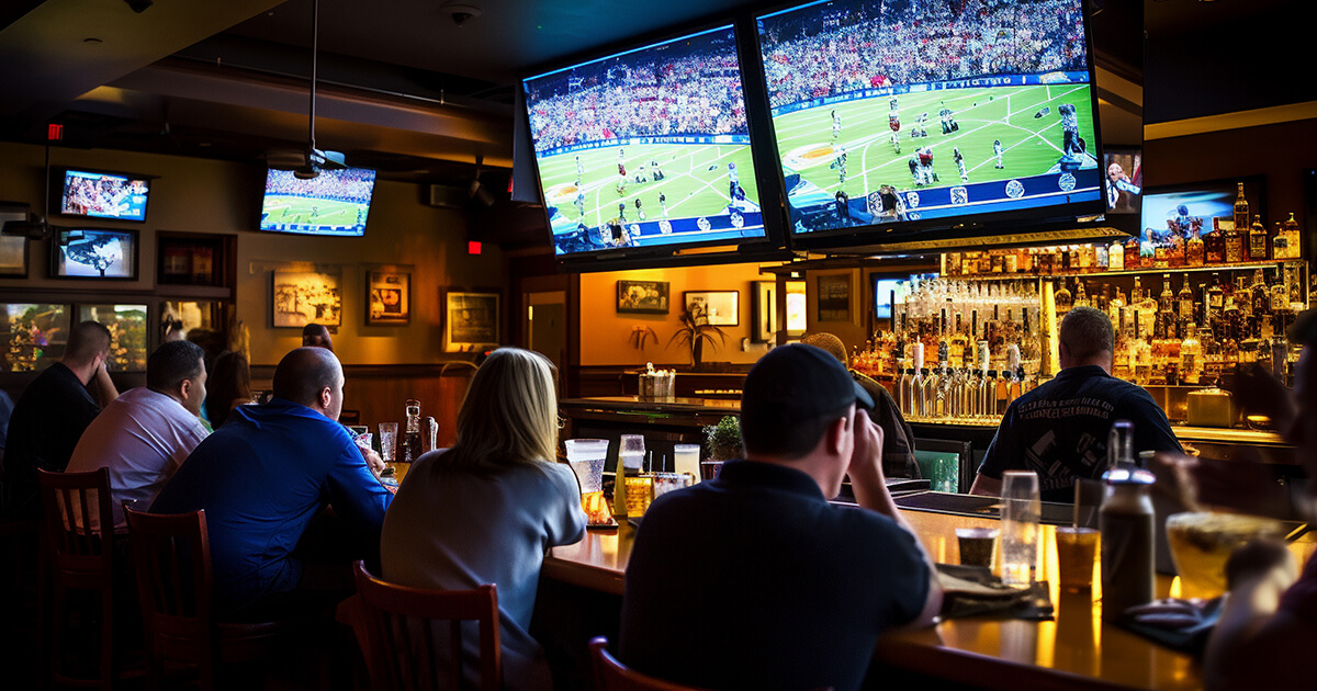 Untitled-1 Patrons in a bar watching a football game on multiple large TVs above the bar counter.