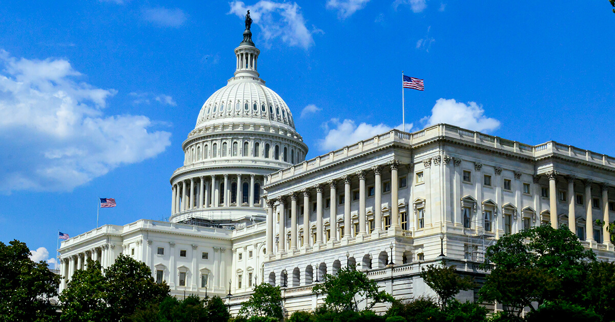 Untitled-1 The United States Capitol building with its large dome and American flags, photographed on a clear day with a blue sky and scattered clouds.