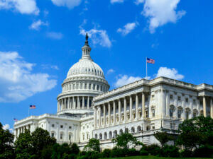 The United States Capitol building with its large dome and American flags, photographed on a clear day with a blue sky and scattered clouds.