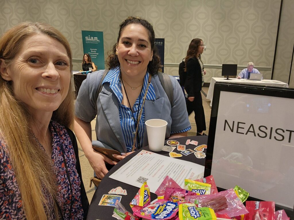 Two NEASIS&T members (also former SLA members) smiling at a small conference table with NEASIST sign, stickers, and candy.