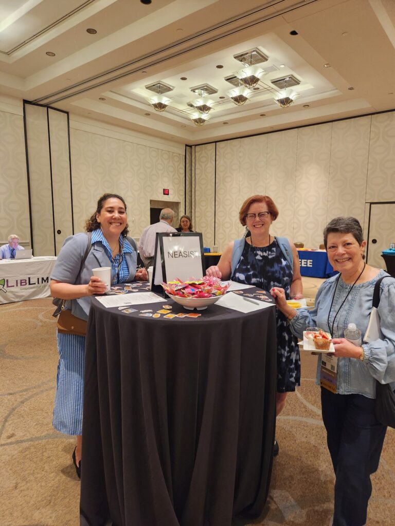 Three conference attendees and members of NEASIS&T standing by a round bar-height table with a NEASIS&T sign.  There are flyers, stickers, and a big bowl of candy.  