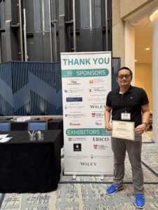Dimaz Ardhi wearing a dark polo shirt and gray pants. He stands next to a thank you sign with the logos of ASIS&T Annual Meeting sponsors.  He holds an award certificate that names his as the recipient of the NEASIST 2025 COnference Support Award.  