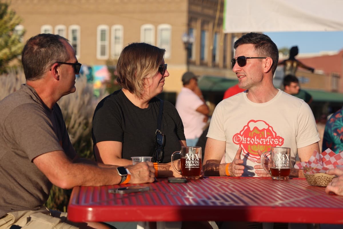 Three people sitting at a table, drinking from steins that read "XIX Brewing company." The text on one individual's shirt reads "Oktoberfest"