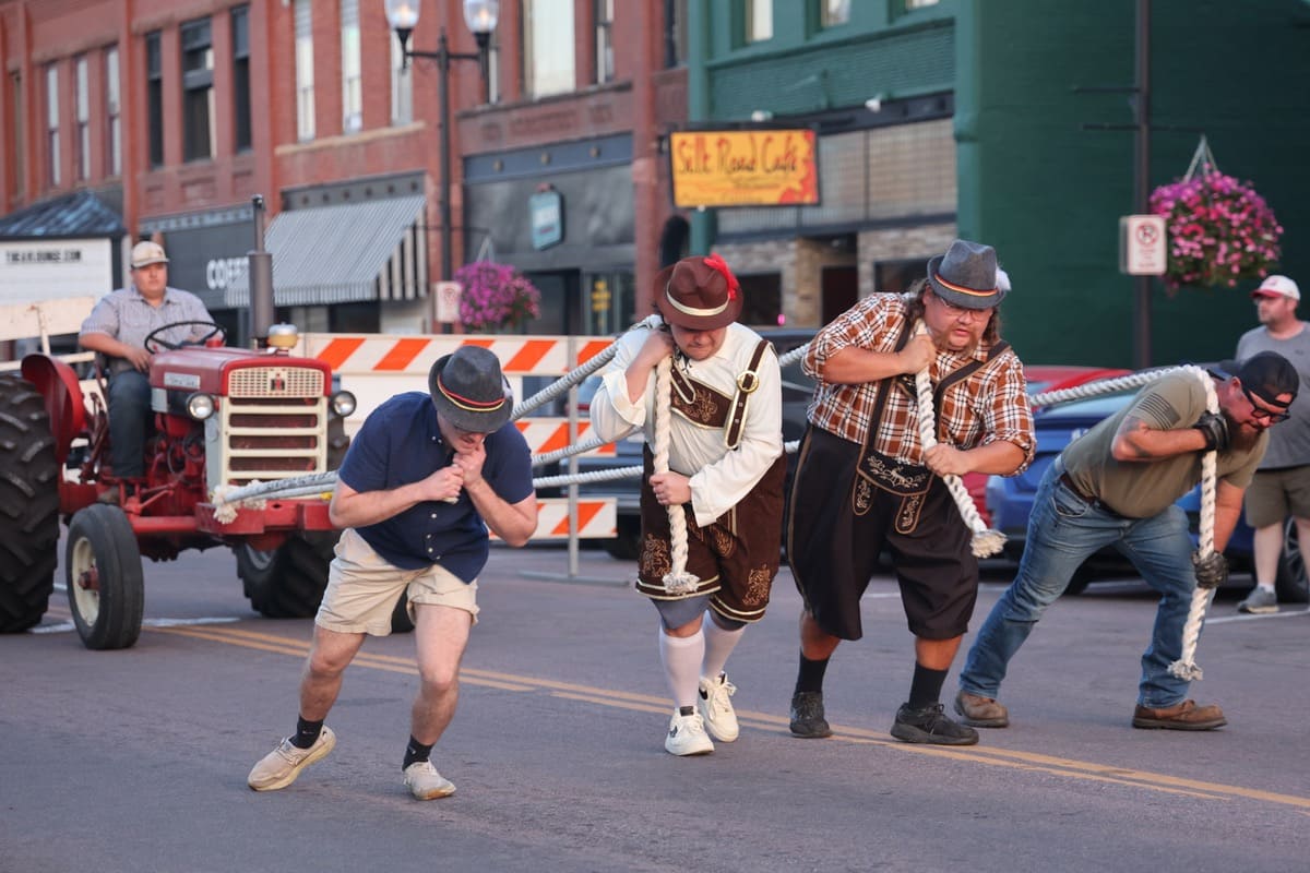 Four people, two of whom are wearing lederhosen, pulling an antique tractor down the street with rope.