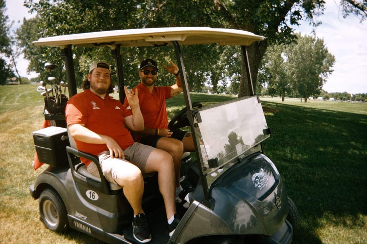 Two people sitting in a golf cart, smiling and making the coyote gesture.
