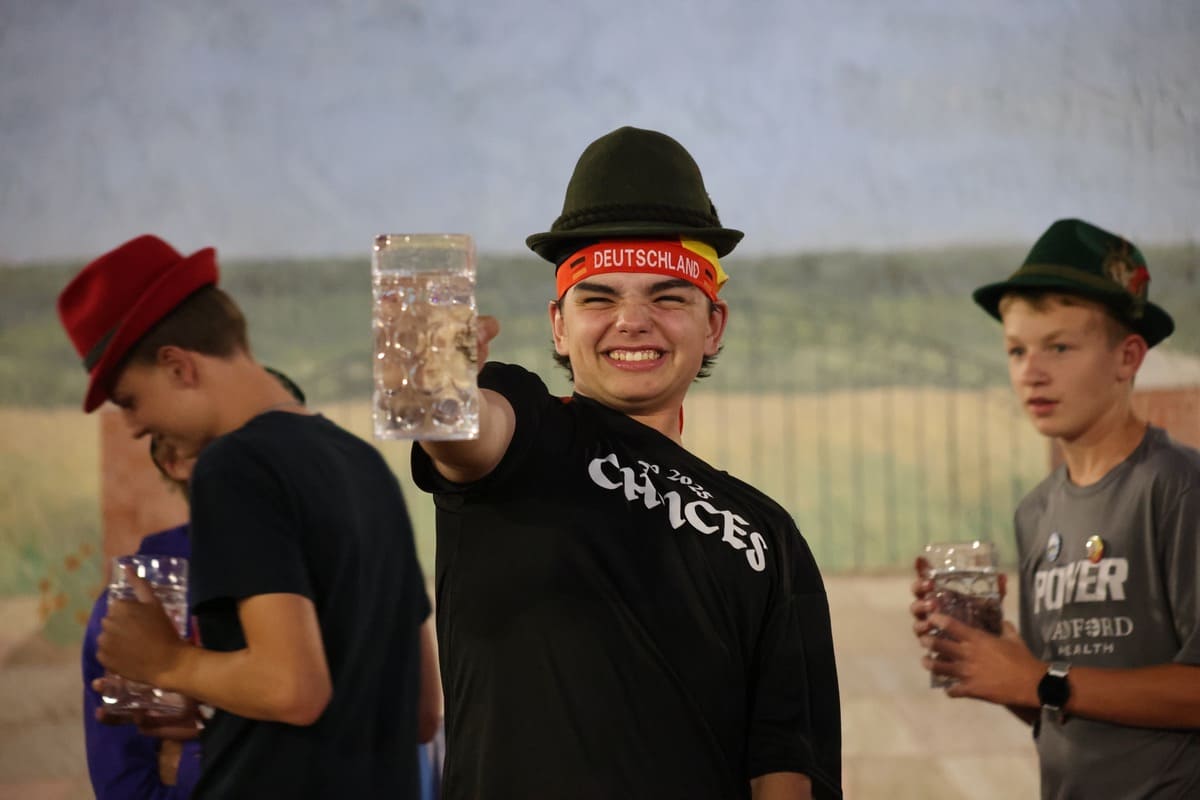 A teenage boy wearing a traditional German hat and headband reading "Deutschland" straining to hold up a stein full of water, while two other boys watch.
