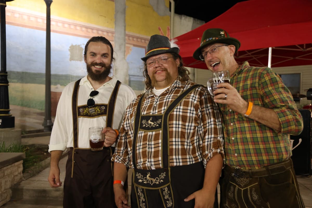 Three people smiling and wearing lederhosen. Two are holding beverages.