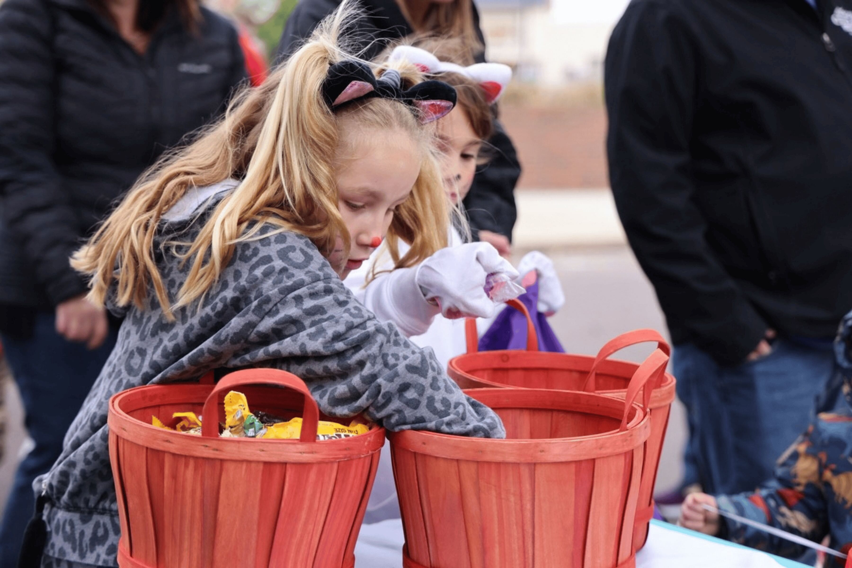 A child dressed as a cat chooses a piece of candy from a basket. Another child, also dressed as a cat, does the same behind them.