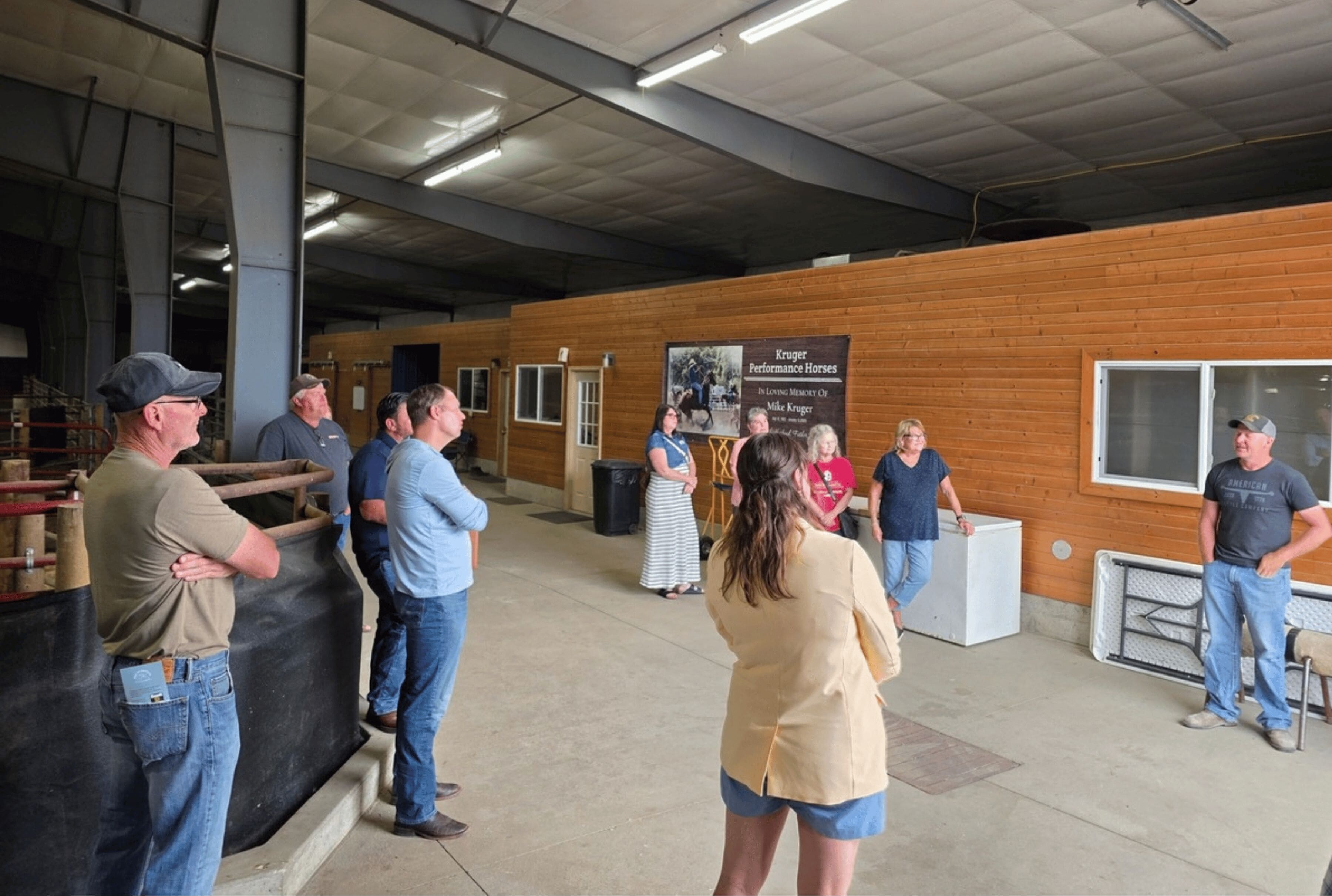 A group of people stand in an agricultural building, listening to someone speak. A banner reading "Kruger Performance Horses" can be seen in the background.