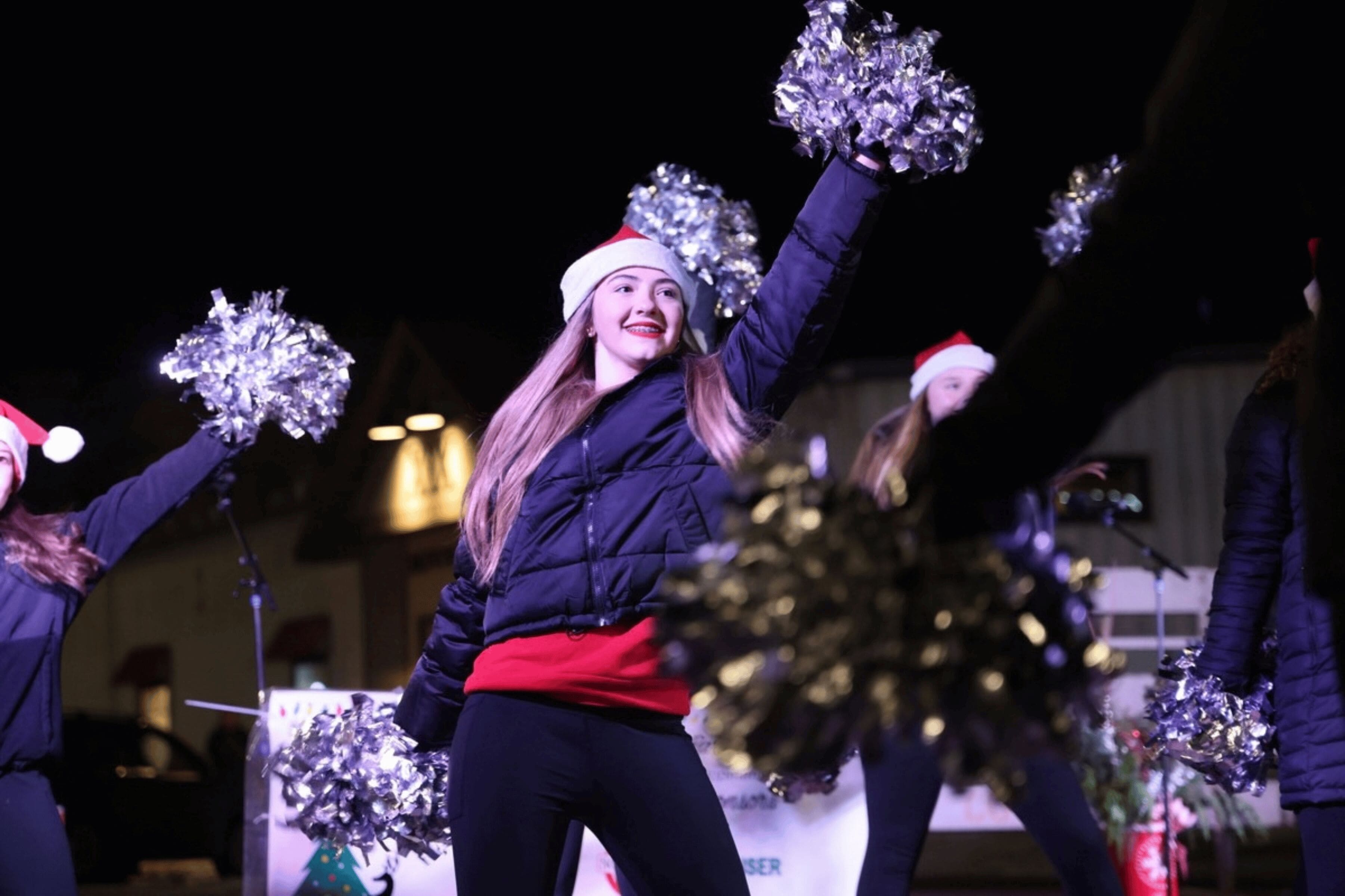 A smiling girl wearing a Santa hat poses with pompoms mid-dance routine.