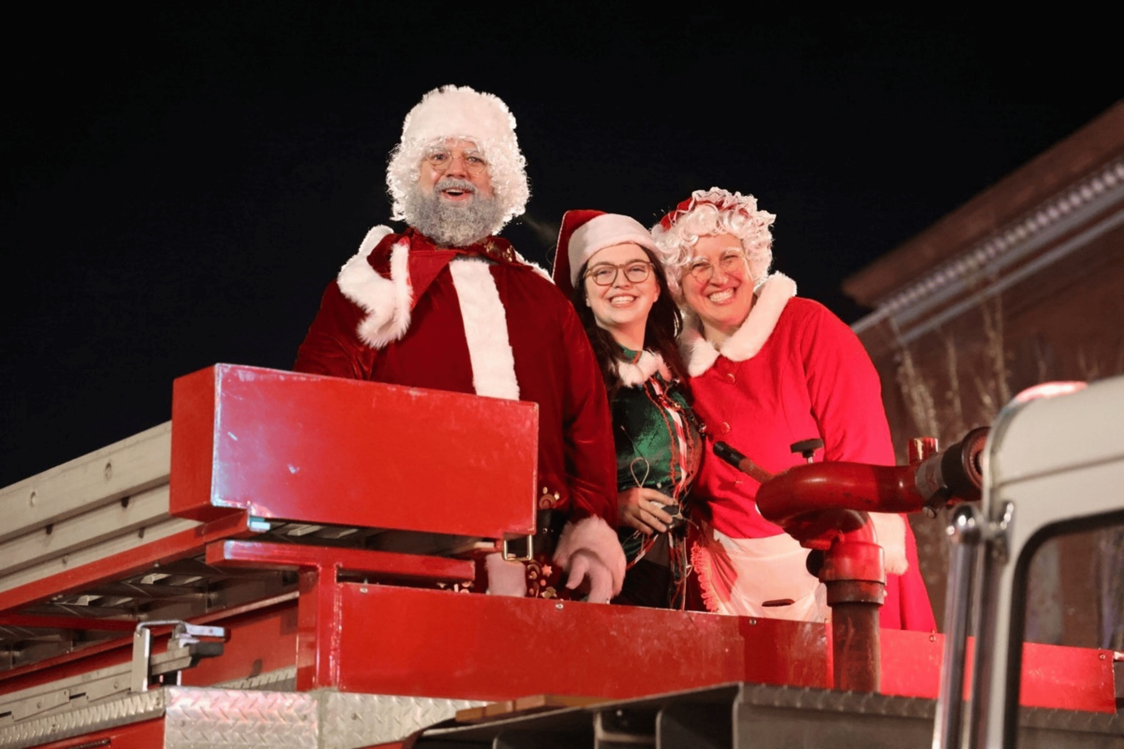 A couple dressed as Santa and Mrs. Claus and a girl dressed as an elf smile atop a firetruck.