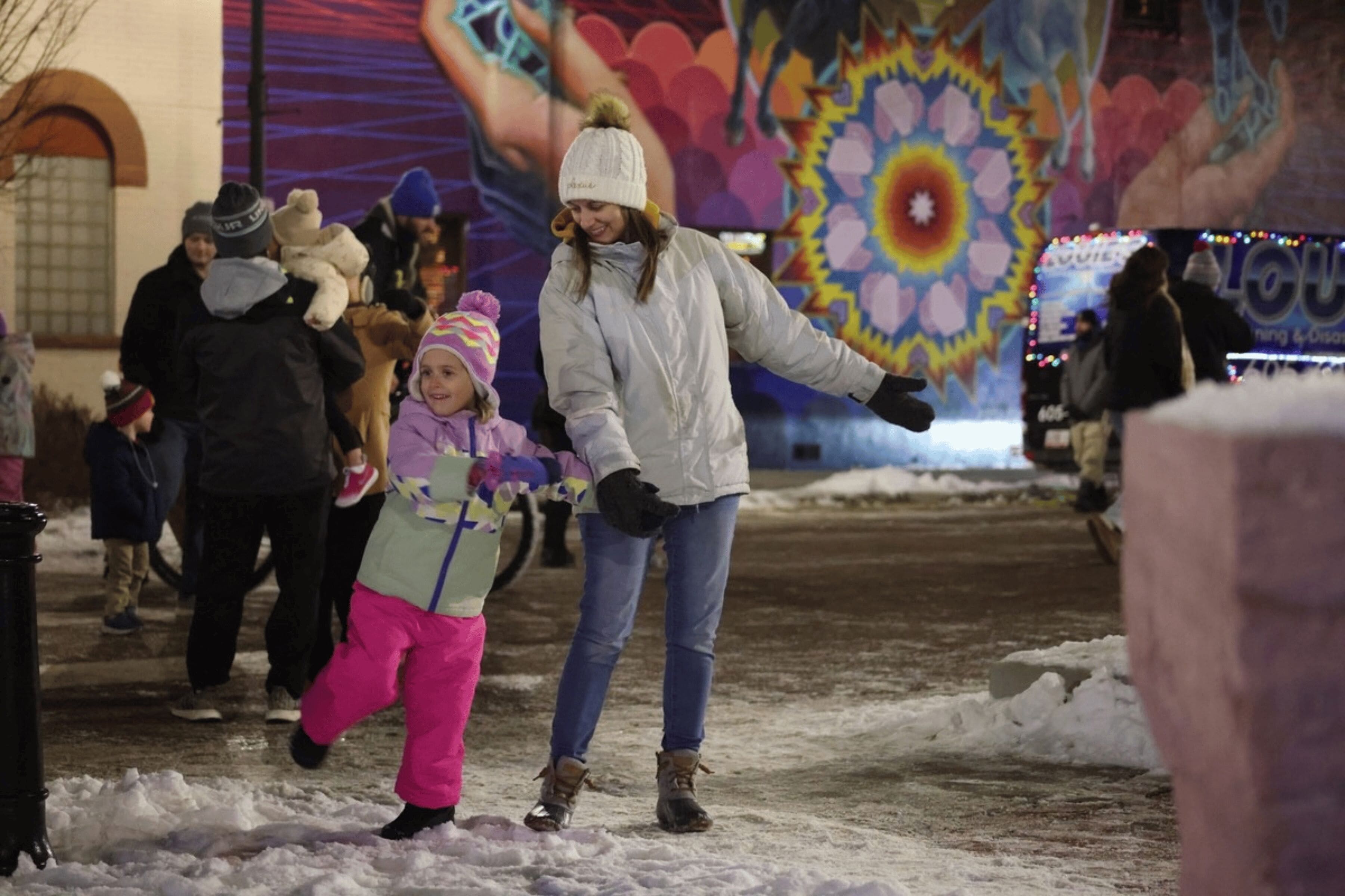 An adult and a child wearing winter clothing hold hands as they dance in the snow.