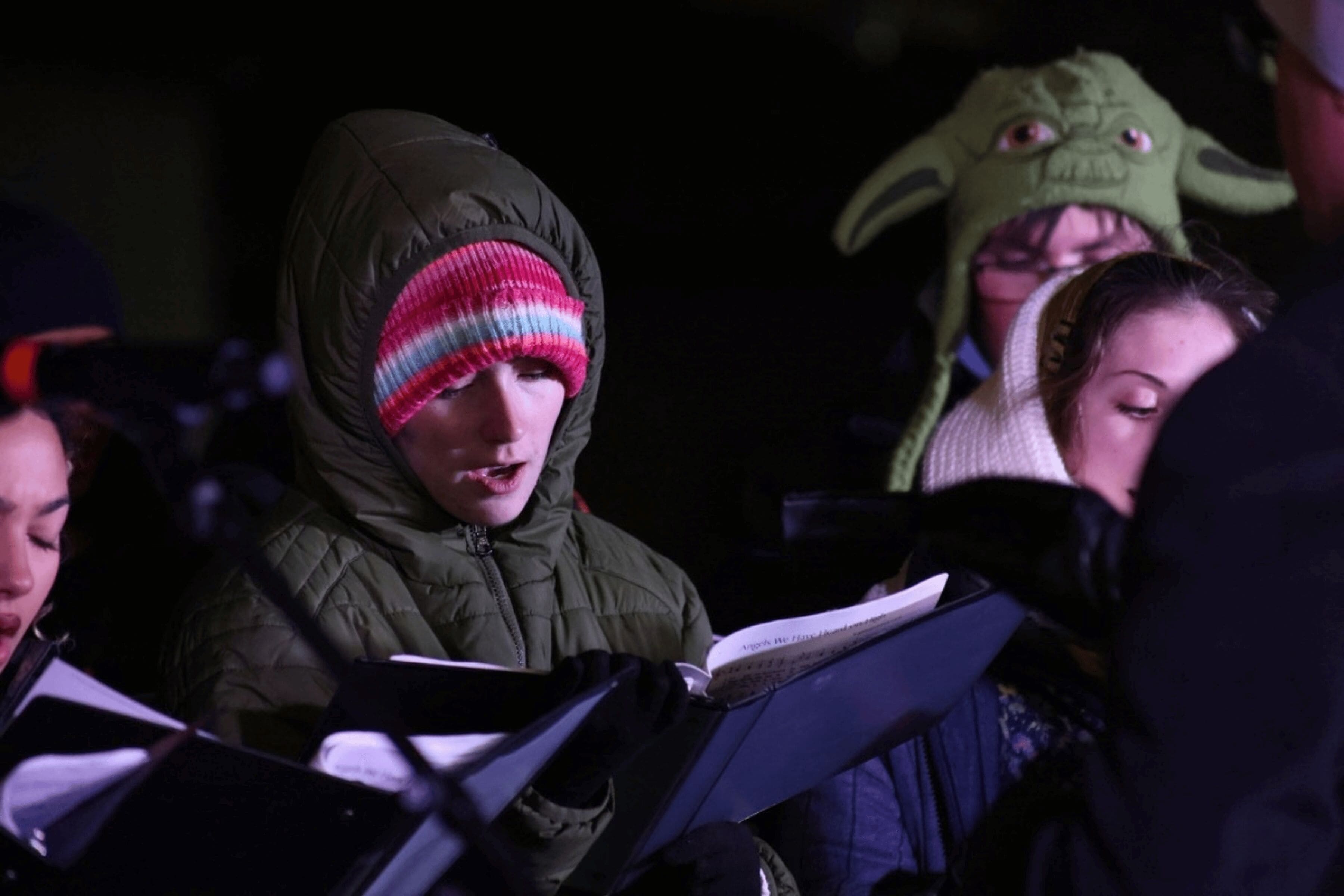 A young woman wearing winter clothing sings carols, surrounded by other choir members.
