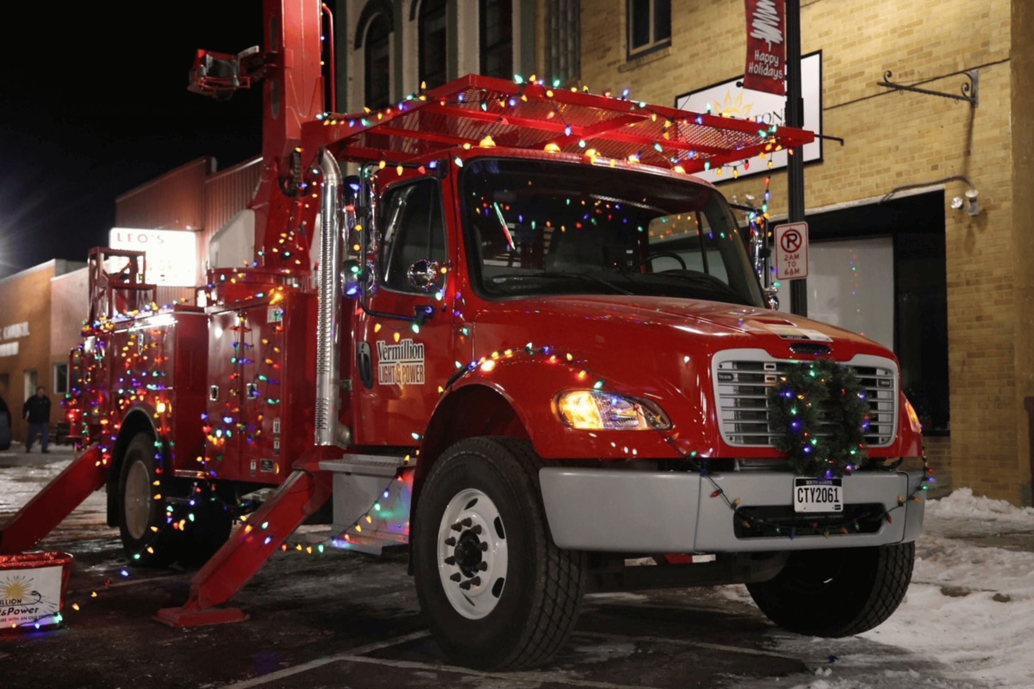 A red industrial truck wrapped in festive lights. The text "Vermillion Light and Power" is visible on the door.