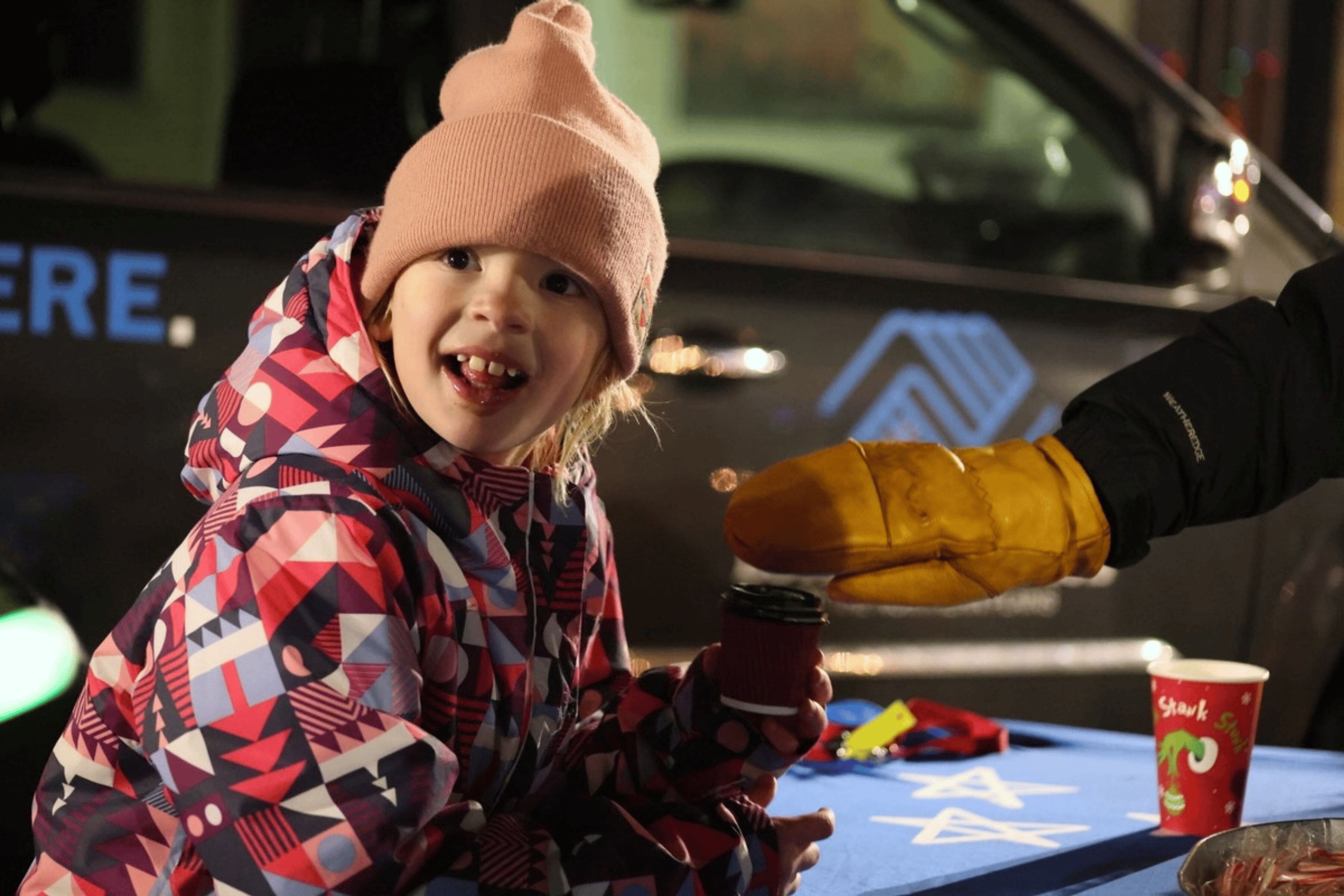 A child is handed a warm beverage by someone wearing mittens.