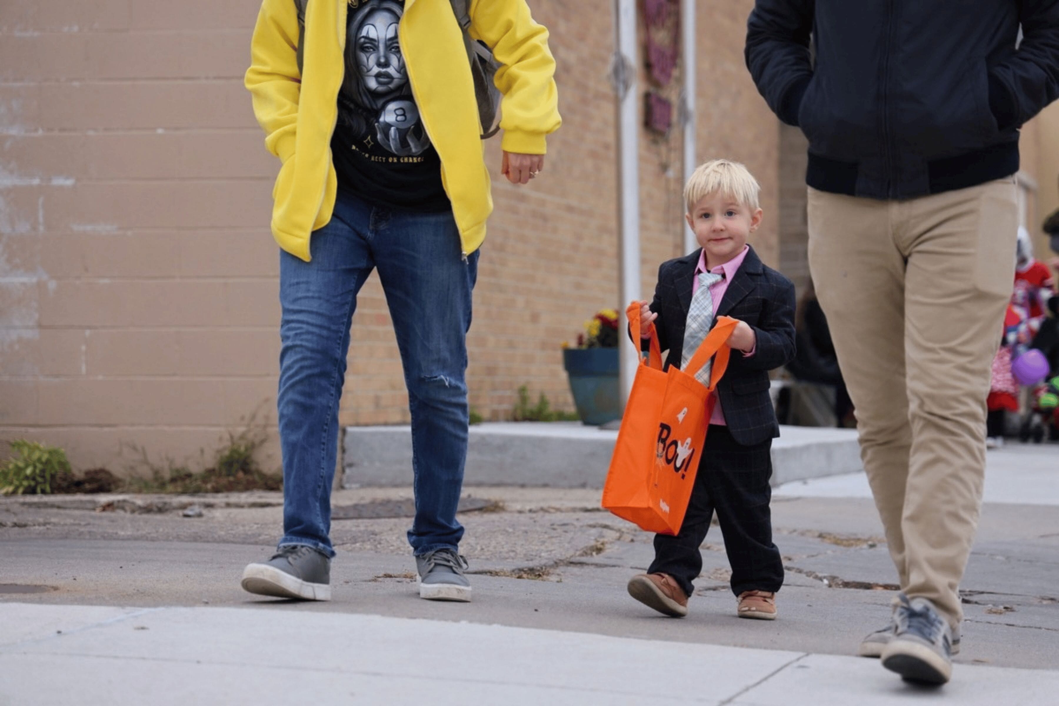 A small child wearing a suit holds an orange bag. The lower bodies of the adults accompanying them can be seen.
