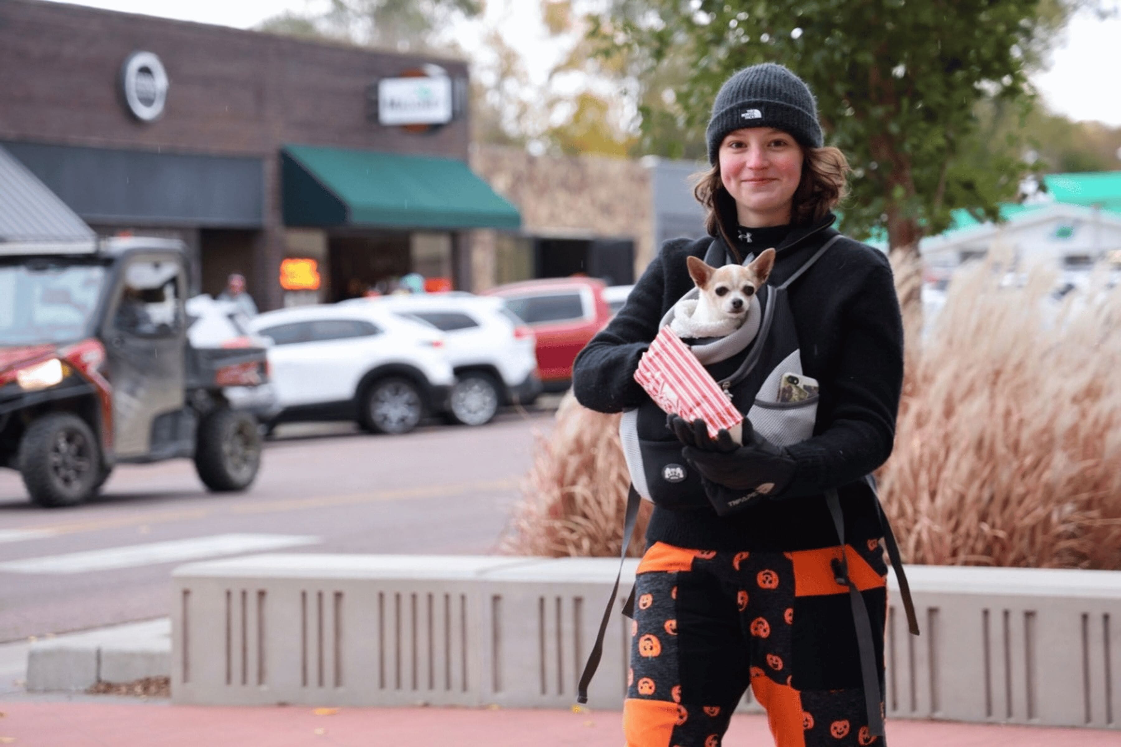 A person wearing pants patterned with Jack-o-Lanterns smiles. There is a small dog in a carrier on their chest.