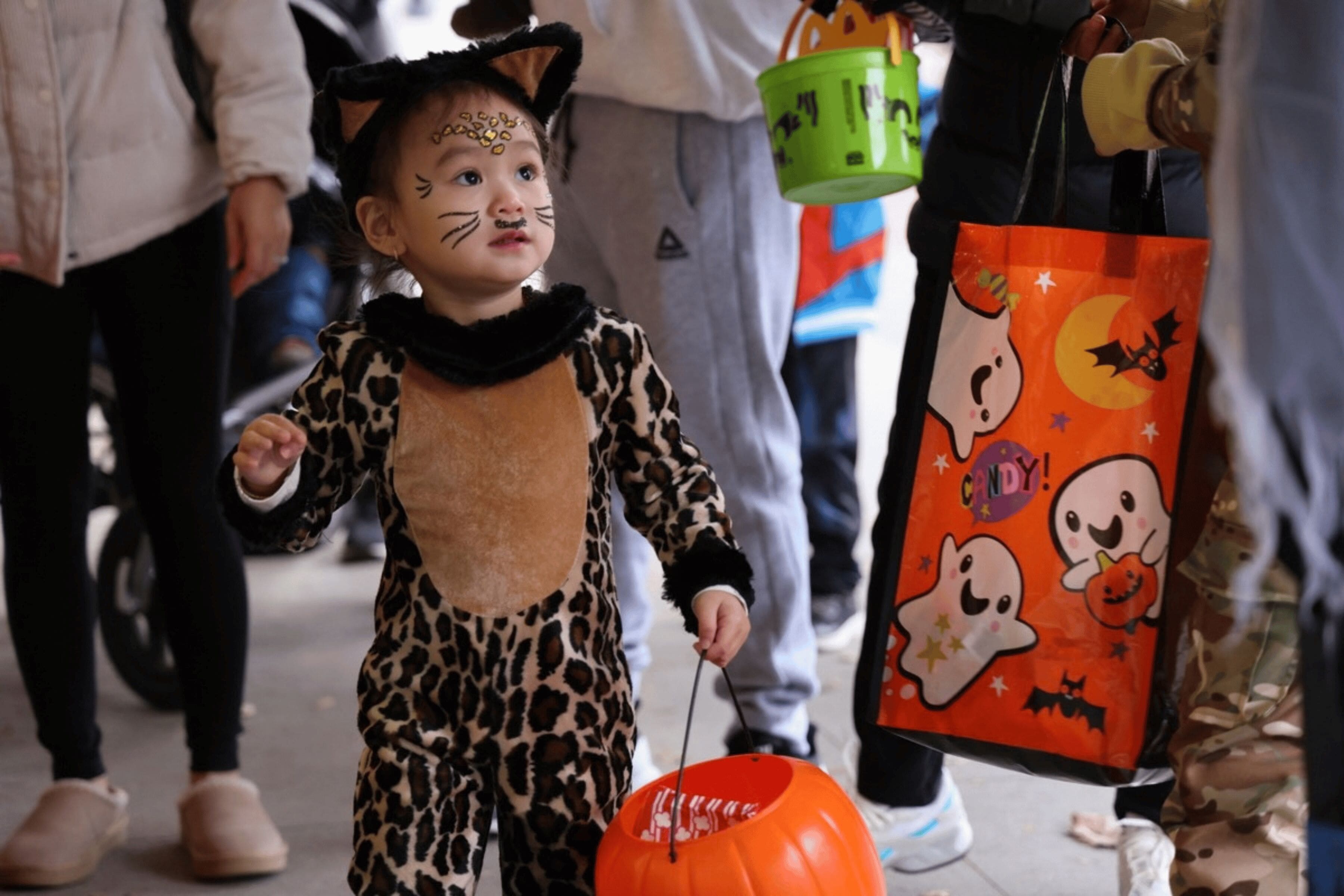 A small child wearing a leopard costume holds a pumpkin-shaped bucket, surrounded by other people.