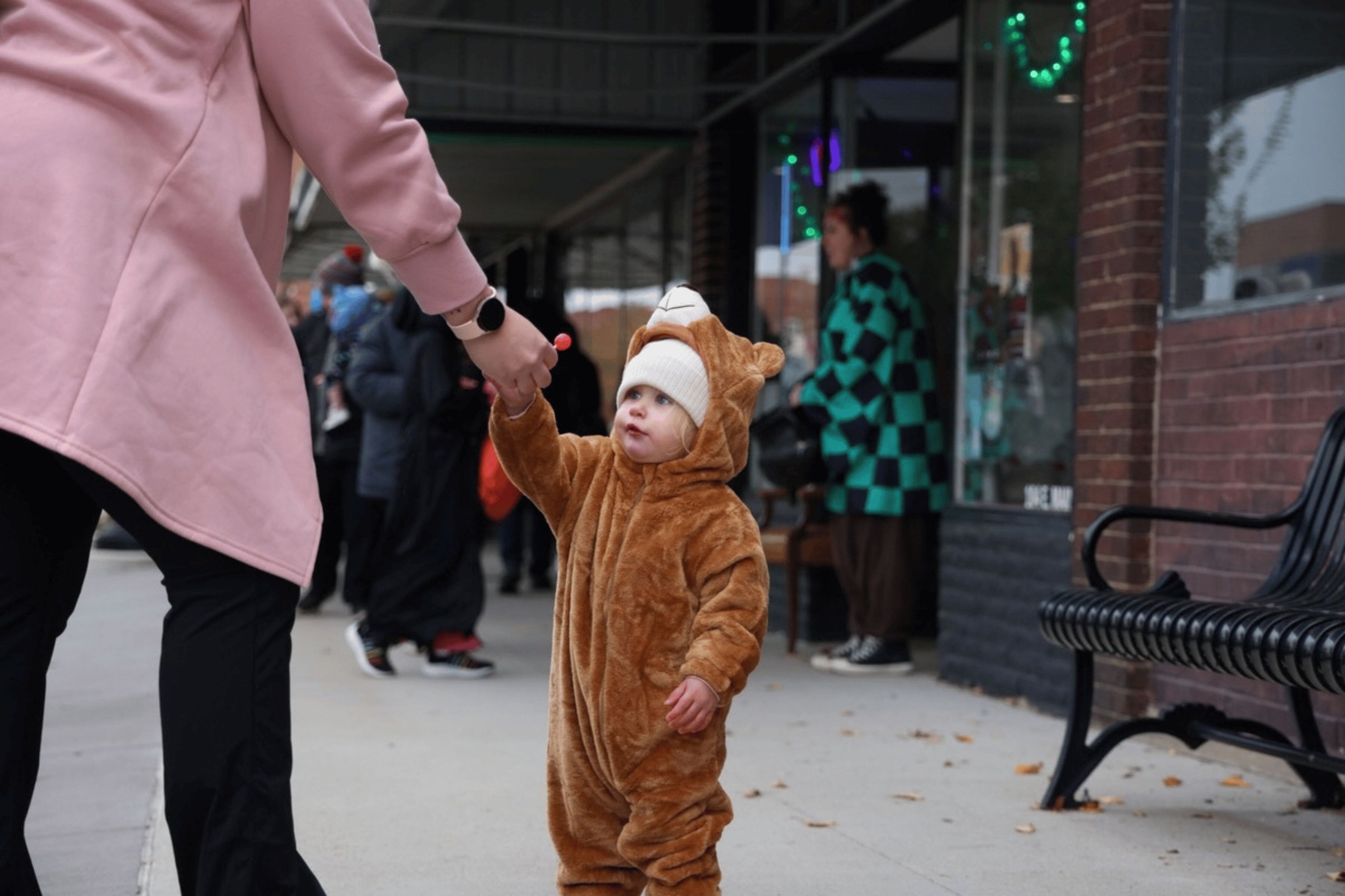 A toddler wearing a bear costume is handed a lollipop by an adult.