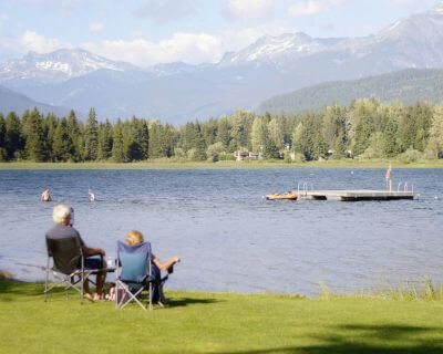 An elderly couple sit in camp chairs on a grassy bank, gazing out on a mountain lake. There is a platform and people enjoying the lake's waters in the distance.