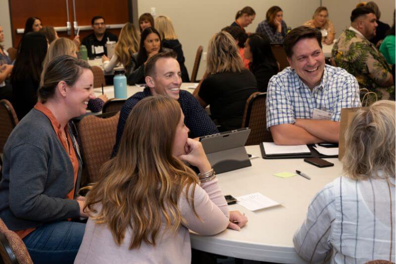 Group of people laughing and talking around a table at a conference.