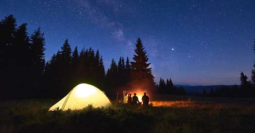 Night camping near fire, forest and mountains on background. Group of friends warming up near bright bonfire. People sitting near tourist illuminated tent under night sky full of stars and milky way.