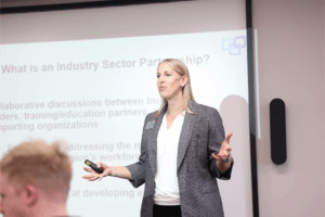Jane Vangsness Frisch discussing workforce talent and attraction at the recent Advanced Manufacturing Industry Sector Partnership meeting. Jane stands in front of the room in front of a projector screen that reads, "What is an industry sector partnership?"