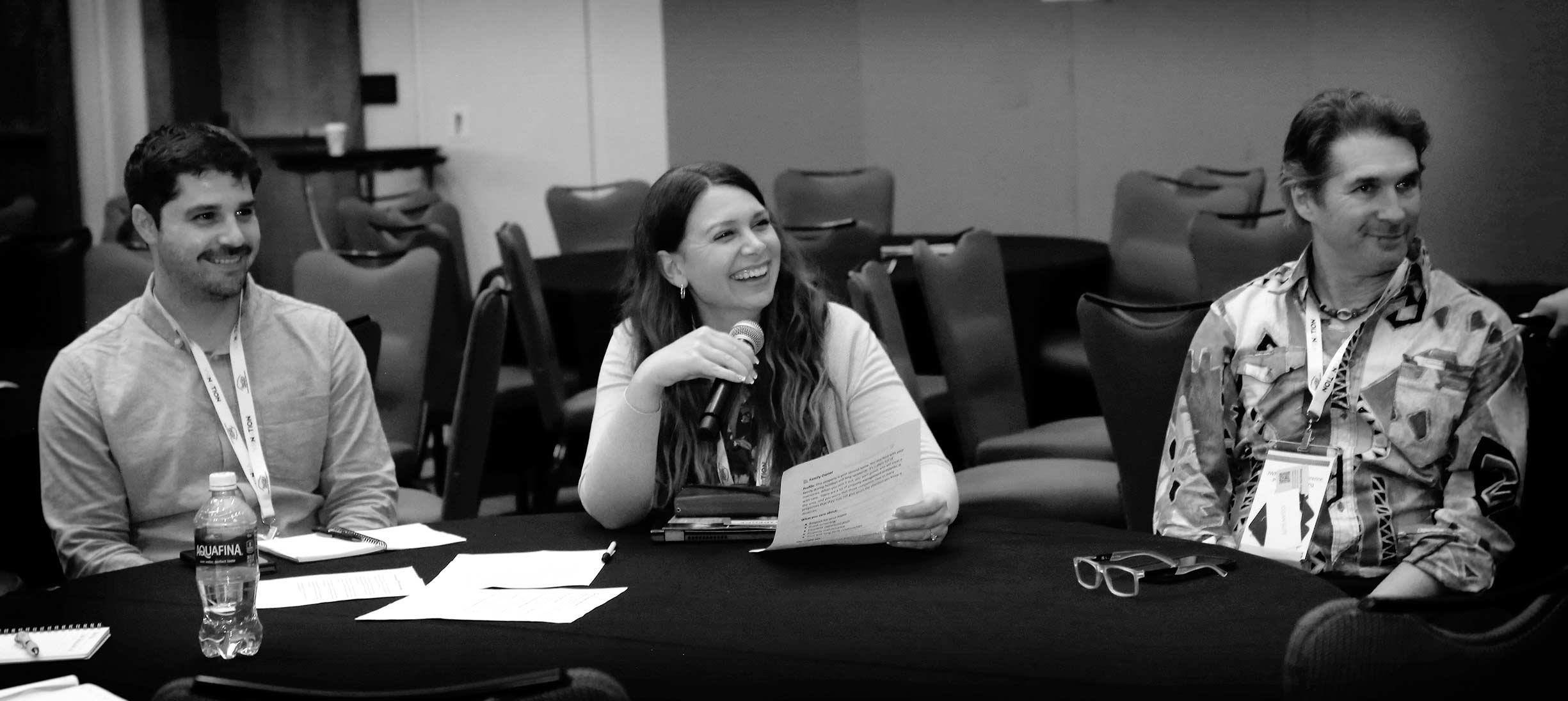 Three conference attendees sit around a table, smiling and talking, while the woman in the center speaks into a microphone.