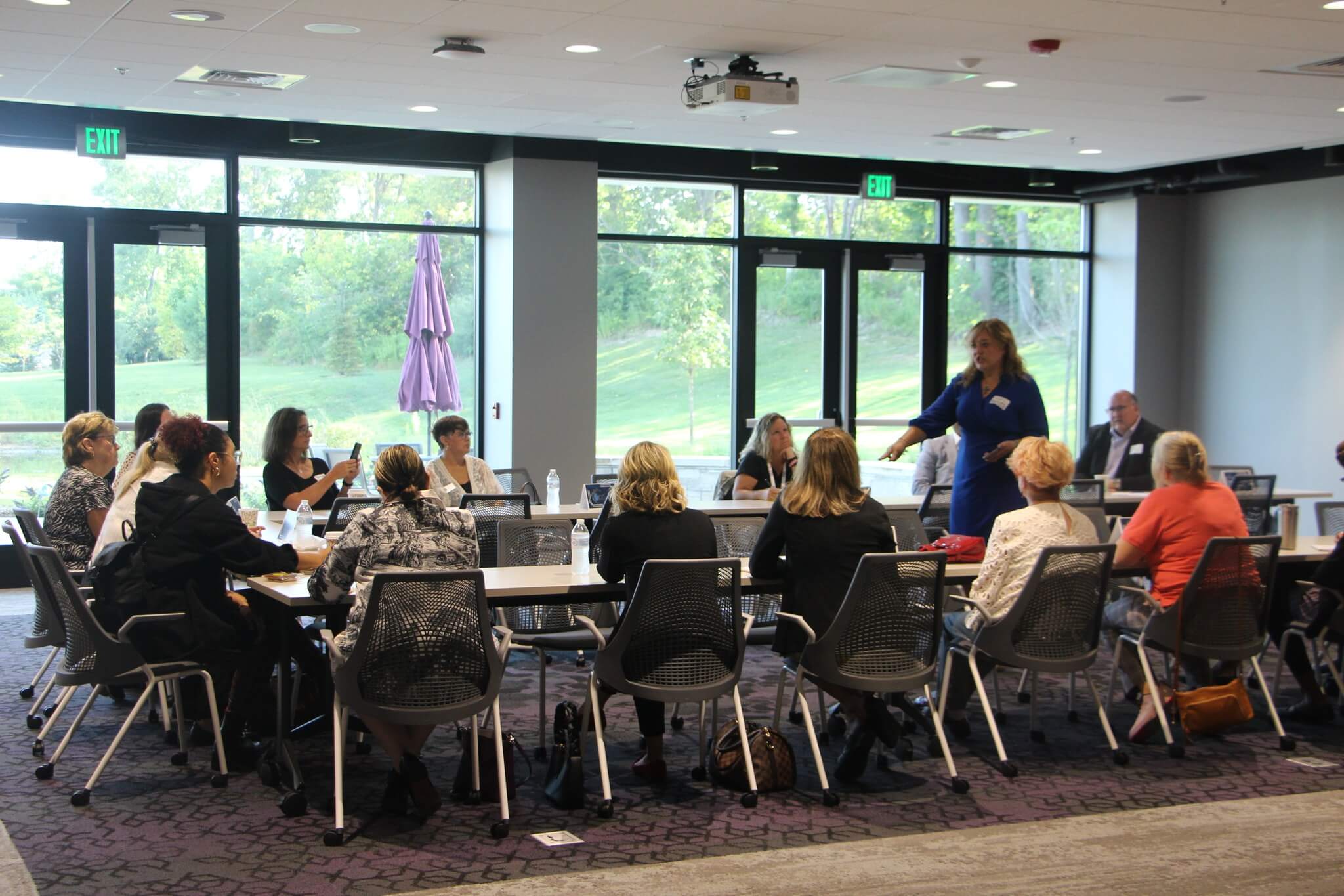 People sit around a conference room table while a woman stands and talks to them.