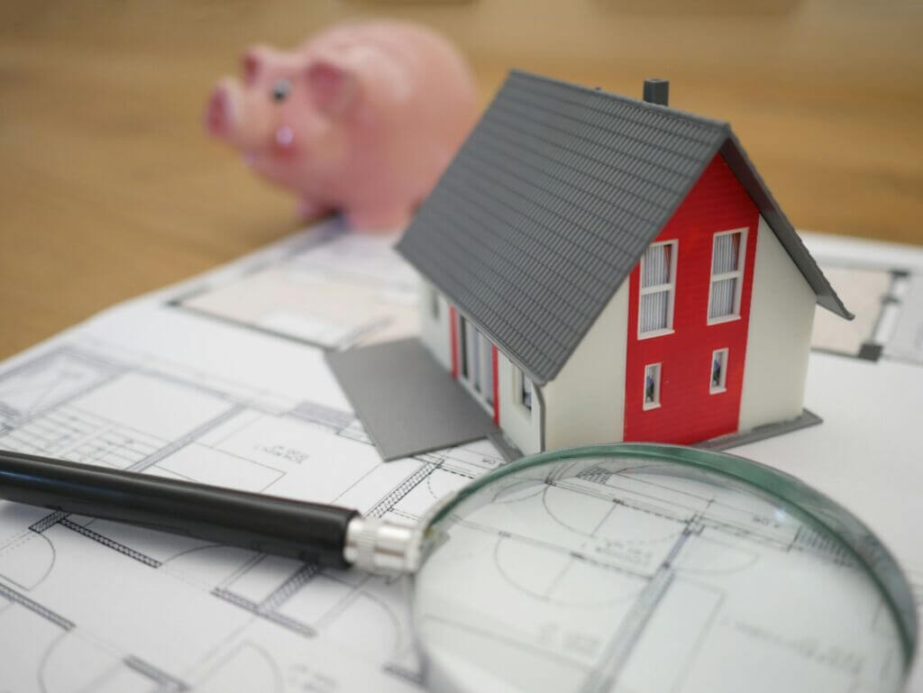 A magnifying glass, house model, and piggy bank sitting on top of house blueprints